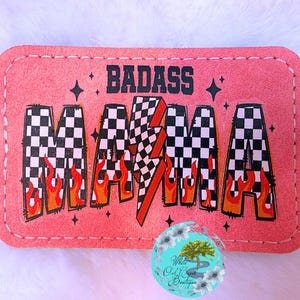 May include: A pink rectangular patch with the words "BADASS MAMA" in black, checkered, and flame-accented lettering. A red lightning bolt is in the center. The patch has a stitched border and small star accents.