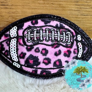 May include: A pink and black leopard print football-shaped patch with white stitching and sequin details. The patch has a black glitter border. A round blue and white logo is in the bottom right corner.