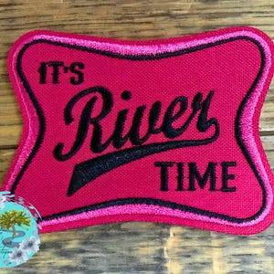May include: A rectangular, pink fabric patch with the words "IT'S River TIME" in black script and block letters. The patch has a black and pink border. A round sticker with a tree and flowers is in the lower left corner.