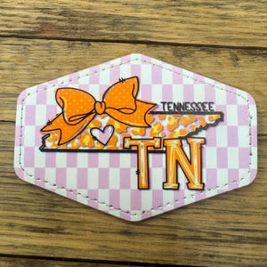 May include: A pink and white checkered hexagon-shaped patch with a large orange bow, a heart, and the outline of the state of Tennessee with the letters "TN" in orange. The text "Tennessee" is written in black above the state outline.