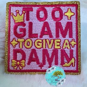 May include: Embroidered patch with the text "TOO GLAM TO GIVE A DAMN" in pink and yellow. The patch features a gold crown, stars, a bow, and a high heel. The patch has a gold glitter border.