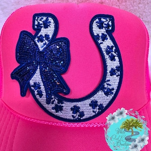 May include: A pink trucker hat with a white and blue horseshoe and a blue sequined bow. The horseshoe has a floral pattern. The hat has a white mesh back.