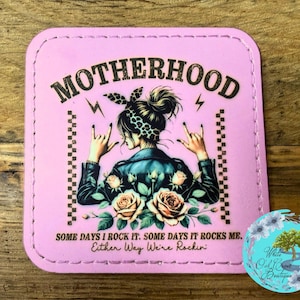 May include: Pink square coaster with the word "MOTHERHOOD" in black. The coaster features a woman with a rock and roll hand gesture, surrounded by roses. Text reads "SOME DAYS I ROCK IT. SOME DAYS IT ROCKS ME. Either Way We're Rockin".