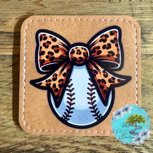 May include: A brown leather coaster with a white baseball with a leopard print bow on top. The coaster has a stitched edge.