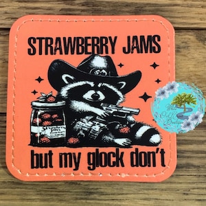 May include: Orange square patch with a black and white cartoon raccoon wearing a cowboy hat and holding a gun. The text "Strawberry Jams but my Glock don't" is printed on the patch.