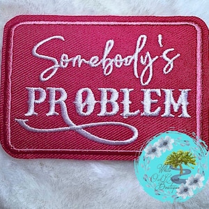 May include: A rectangular, embroidered patch in a vibrant fuchsia color. The patch features the words "Somebody's PROBLEM" in white script and block letters. A circular logo with a tree and flowers is in the lower right corner.