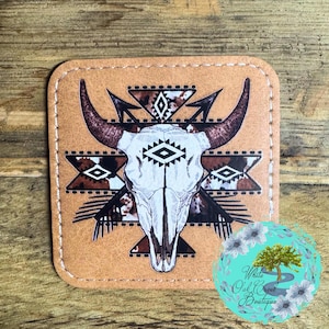 May include: A brown leather coaster with a white bull skull and geometric pattern. The skull is surrounded by a black and brown geometric pattern with arrows pointing towards the skull. The coaster has a stitched edge.