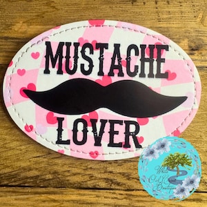 May include: Oval-shaped patch with the words "MUSTACHE LOVER" in black letters, a black mustache graphic, and a pink and white checkered background with pink hearts. The patch has a white stitched border.