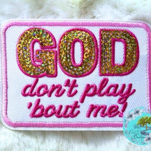 May include: A rectangular embroidered patch with a white background and a pink border. The word GOD is featured in large, gold, sequined letters outlined in pink. Below, the text don't play 'bout me! is embroidered in pink thread.