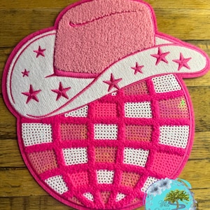 May include: A pink and white felt patch with a pink cowboy hat and a pink and white checkered design. The patch has white stars around the brim of the hat.