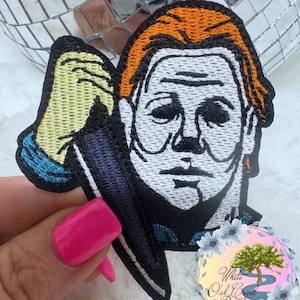 May include: An embroidered iron-on patch featuring a white-faced character with orange hair holding a knife. The character has a menacing expression and is wearing a blue and white shirt. The patch is perfect for adding a spooky touch to clothing, bags, or other accessories.