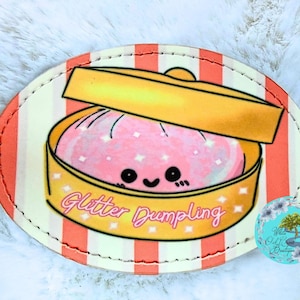 May include: An oval-shaped patch featuring a cartoon dumpling in a yellow steamer basket. The pink dumpling has a smiling face and sparkles. The text "Glitter Dumpling" is written in cursive below the basket. The background has orange and white stripes.