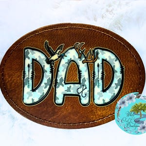 May include: A brown, oval-shaped patch with the word "DAD" in a camouflage pattern. The letters are decorated with hunting-themed elements, including antlers, arrows, and a duck. The patch has a stitched border and a small, round logo in the bottom right corner.