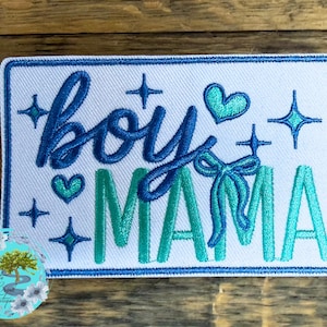 May include: A rectangular patch with a white background and a blue border. The patch features the words "boy MAMA" in blue and teal script, along with teal hearts, stars, and a bow. The patch is likely an embroidered item.