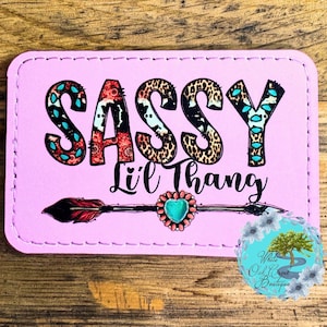 May include: A pink rectangular patch with the words "SASSY Lil' Thang" in a mix of fonts and patterns. The "S" and "Y" are turquoise, the "A" is red floral, and the "S"s are leopard print. An arrow with a turquoise heart is below the text.