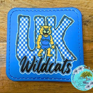 May include: A blue square patch featuring the letters "UK" in a checkered pattern, with a cartoon wildcat mascot in the center. The word "Wildcats" is written in a script font below the letters. The patch is on a wooden surface.