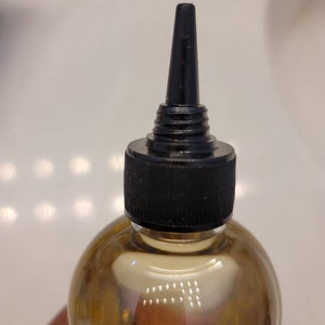 Puede incluir: Una botella de vidrio transparente con una tapa negra y un pico largo y delgado. La botella est&aacute; llena de un l&iacute;quido marr&oacute;n claro.