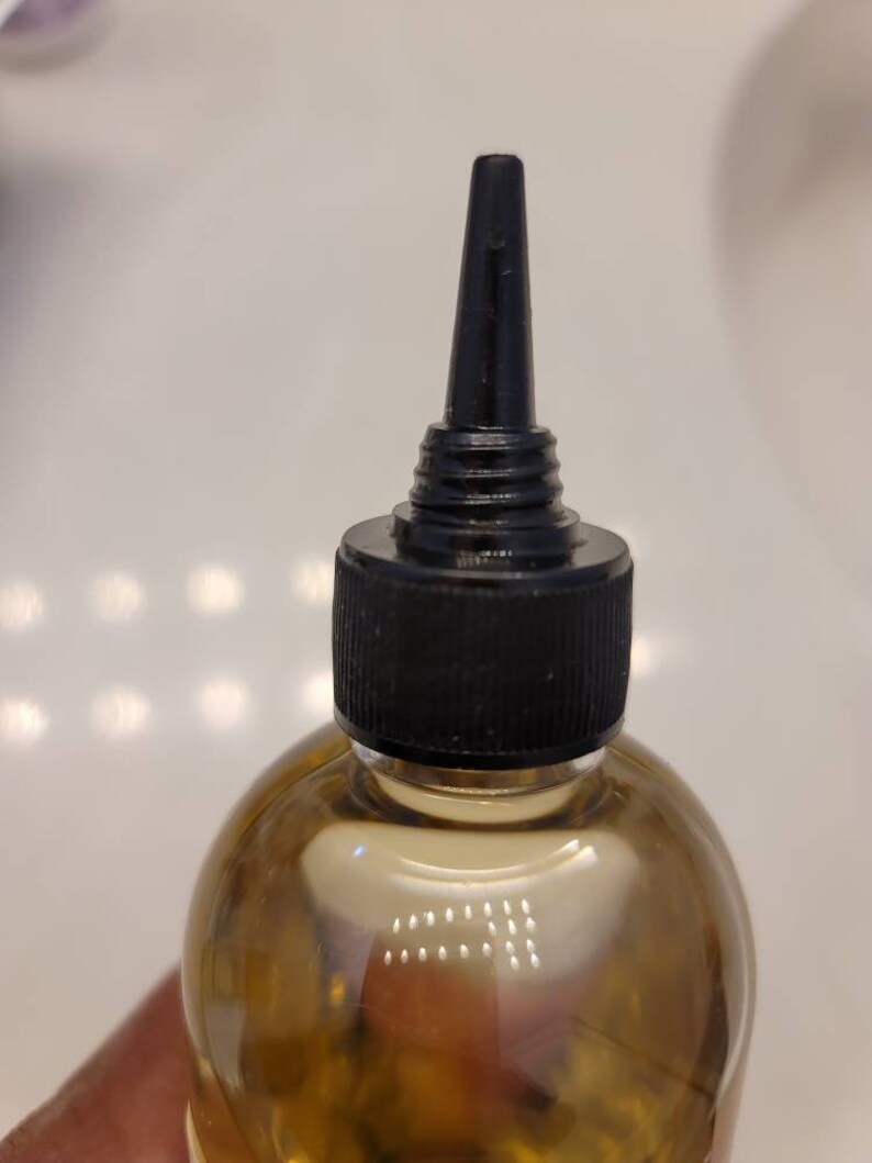 Puede incluir: Una botella de vidrio transparente con una tapa negra y un pico largo y delgado. La botella est&aacute; llena de un l&iacute;quido marr&oacute;n claro.