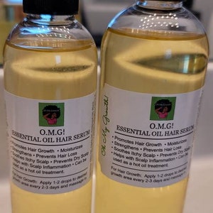 Puede incluir: Dos botellas de vidrio transparente con tapas negras y etiquetas que dicen "O.M.G! Suero capilar de aceite esencial". Las etiquetas enumeran los beneficios del suero, incluyendo la promoci&oacute;n del crecimiento del cabello, la hidrataci&oacute;n y la prevenci&oacute;n de la p&eacute;rdida de cabello. Las botellas est&aacute;n sobre una superficie blanca.
