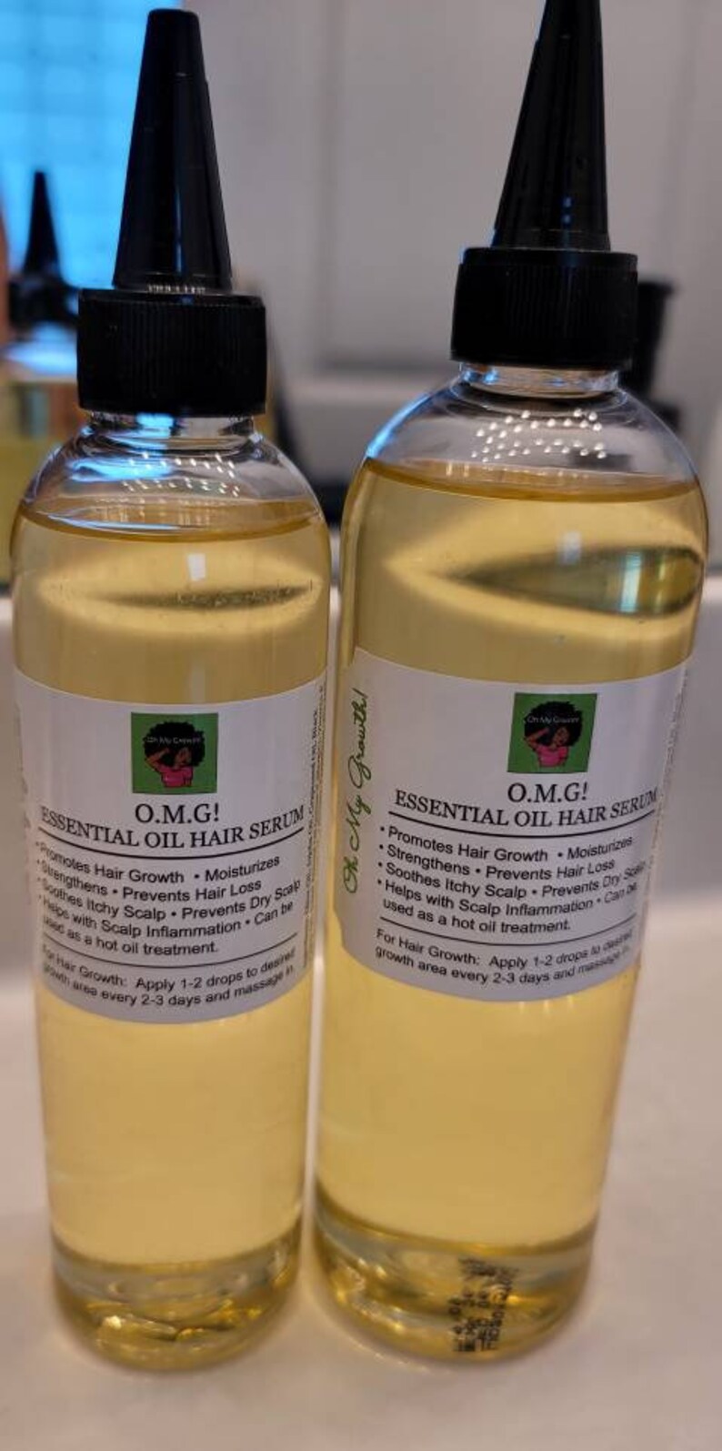 Puede incluir: Dos botellas de vidrio transparente con tapas negras y etiquetas que dicen "O.M.G! Suero capilar de aceite esencial". Las etiquetas enumeran los beneficios del suero, incluyendo la promoci&oacute;n del crecimiento del cabello, la hidrataci&oacute;n y la prevenci&oacute;n de la p&eacute;rdida de cabello. Las botellas est&aacute;n sobre una superficie blanca.