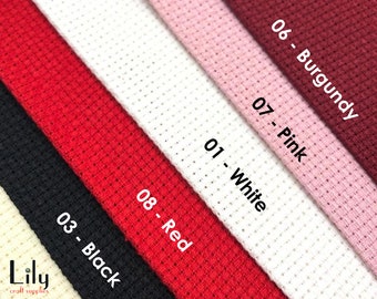 May include: A close-up of eight different colours of Aida cloth, a type of fabric used for cross-stitch embroidery. The colours are white, pink, burgundy, red, black, and three shades of beige. The fabric is labelled with numbers and colour names.