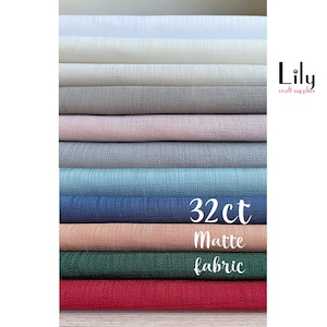 Puede incluir: Una pila de 10 colores diferentes de tejido mate de 32 hilos. Los colores son blanco, blanco roto, gris, rosa claro, azul claro, azul, melocotón, verde y rojo. El tejido está etiquetado como "32ct Matte Fabric".