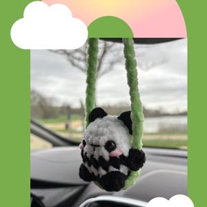 Może przedstawiać: Ręcznie robiony szydełkowy urok do samochodu z pandą. Panda ma czarno-biały wzór z różowymi policzkami i czarnymi uszami. Urok jest zawieszony na zielonej włóczce. Tło zawiera tekst "Crochet Panda".