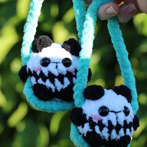 May include: Two handmade, plush panda bear purses with turquoise straps. The pandas have black and white fur, black eyes, and pink cheeks. The purses are held against a blurred green background.