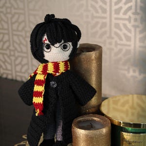 May include: A crocheted doll of a young boy with black hair, wearing a black coat, a red and yellow scarf, and round glasses. The doll is standing on a gold tray with candles.