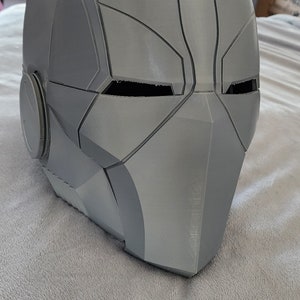 Könnte beinhalten: Ein grauer 3D-gedruckter Helm mit futuristischem Design. Der Helm hat ein Visier und ein rundes, graues Detail an der Seite.