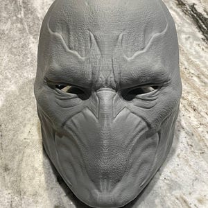 May include: A gray, textured mask with a detailed, alien-like design. The mask features prominent brow ridges, defined cheekbones, and a sculpted face. The eye openings are visible, and the overall aesthetic suggests a futuristic or science fiction theme.