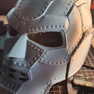 Può includere: Un casco grigio stampato in 3D con un design dettagliato, che presenta una superficie testurizzata e numerose piccole indentature circolari. Il casco è appoggiato su una superficie grigliata nera con un righello e un calibro digitale.