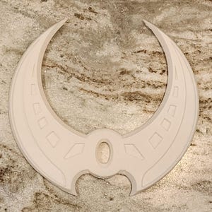 以下が含まれることがあります： 幾何学模様の白い三日月型の装飾品。デザインは中央の楕円と、湾曲したエッジに沿ったダイヤモンドの形が特徴です。このオブジェクトは、装飾要素またはより大きなデザインの一部である可能性があります。