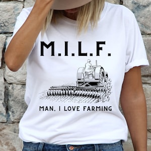 Op de afbeelding: Wit T-shirt met de tekst "M.I.L.F. Man, I Love Farming" en een zwart-witte illustratie van een tractor.