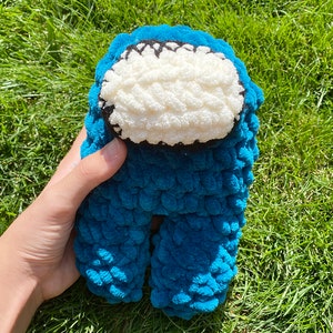 May include: A blue crocheted plush toy shaped like the character from the video game Among Us. The toy has a white face with a black outline.