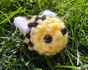 Peut inclure: Un jouet d'abeille au crochet fait à la main, avec un corps jaune, des rayures noires et des ailes blanches. L'abeille a de grands yeux noirs et brillants. Le jouet est posé dans l'herbe verte.