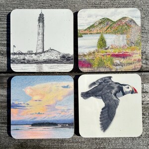 May include: Set of four square coasters featuring nature scenes. One coaster shows a lighthouse on a rocky shore, another shows a lake with a mountain in the background, a third shows a sunset over the water, and the last shows a puffin in flight.