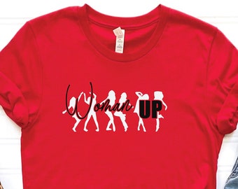 Pode incluir: Camiseta vermelha com um gráfico branco de mulheres em silhueta com o texto "Woman Up" em preto.