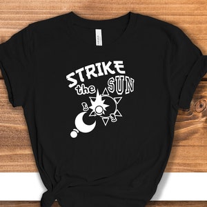Könnte beinhalten: Schwarzes T-Shirt mit weißem Text "Strike the Sun" und einer Grafik von Sonne und Mond.