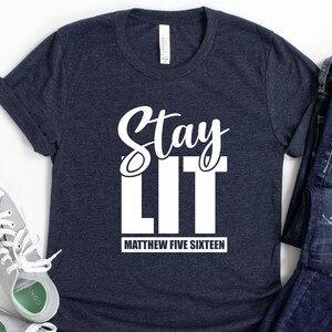 以下が含まれることがあります： 白い文字で「Stay Lit」と「Matthew Five Sixteen」と書かれたネイビーブルーのTシャツです。