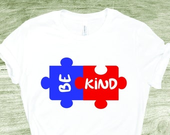 May include: White t-shirt with a blue and red puzzle piece design. The puzzle pieces are joined together and say "BE KIND" in white text.