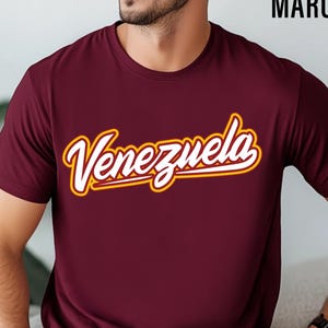 May include: Maroon t-shirt with the word "Venezuela" in a white script font with a gold outline. The shirt is short-sleeved and made of a soft material.