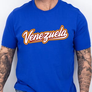 May include: Royal blue t-shirt with the word "Venezuela" in a cursive, yellow and red font. The shirt is a solid color and has short sleeves. The text "ROYAL BLUE" is in black, block letters.