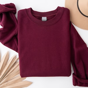 Puede incluir: Una sudadera de cuello redondo y manga larga de color burdeos. La sudadera est&aacute; colocada sobre una superficie blanca con una hoja de palma y un sombrero marr&oacute;n.