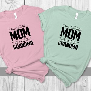 May include: Two t-shirts with the text "I have two titles MOM and GRANDMA" in black on a pink and a mint green shirt.