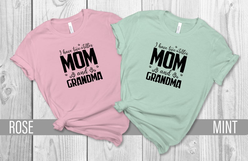May include: Two t-shirts with the text "I have two titles MOM and GRANDMA" in black on a pink and a mint green shirt.