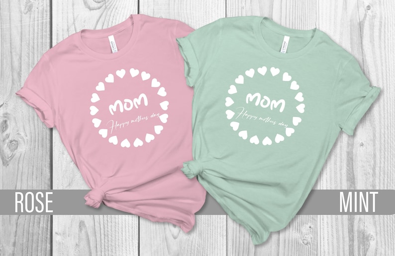 May include: Two t-shirts with a white circle of hearts around the word "MOM" and the text "Happy Mother's Day". The t-shirt on the left is pink and the t-shirt on the right is mint green.