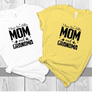 May include: Two t-shirts, one white and one yellow, with the text "I have two titles MOM and GRANDMA" printed in black. The text is surrounded by small paw prints and hearts.