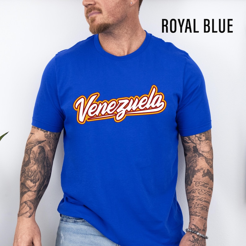May include: Royal blue t-shirt with the word "Venezuela" in a cursive, yellow and red font. The shirt is a solid color and has short sleeves. The text "ROYAL BLUE" is in black, block letters.