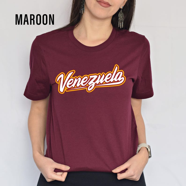 May include: Maroon crew neck t-shirt with the word "Venezuela" in a white and yellow script. The word "MAROON" is printed in the upper left corner of the image.